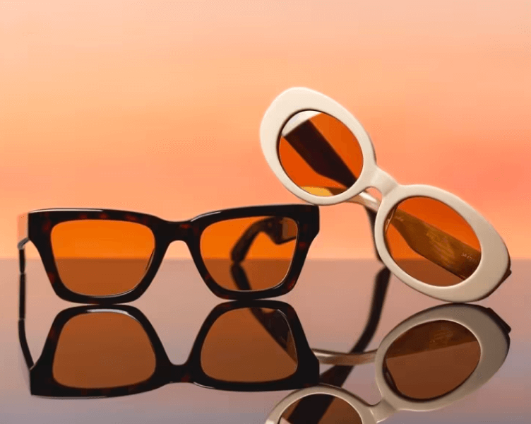 Two pairs of retro sunglasses with amber lenses reflected on glossy surface against coral backdrop