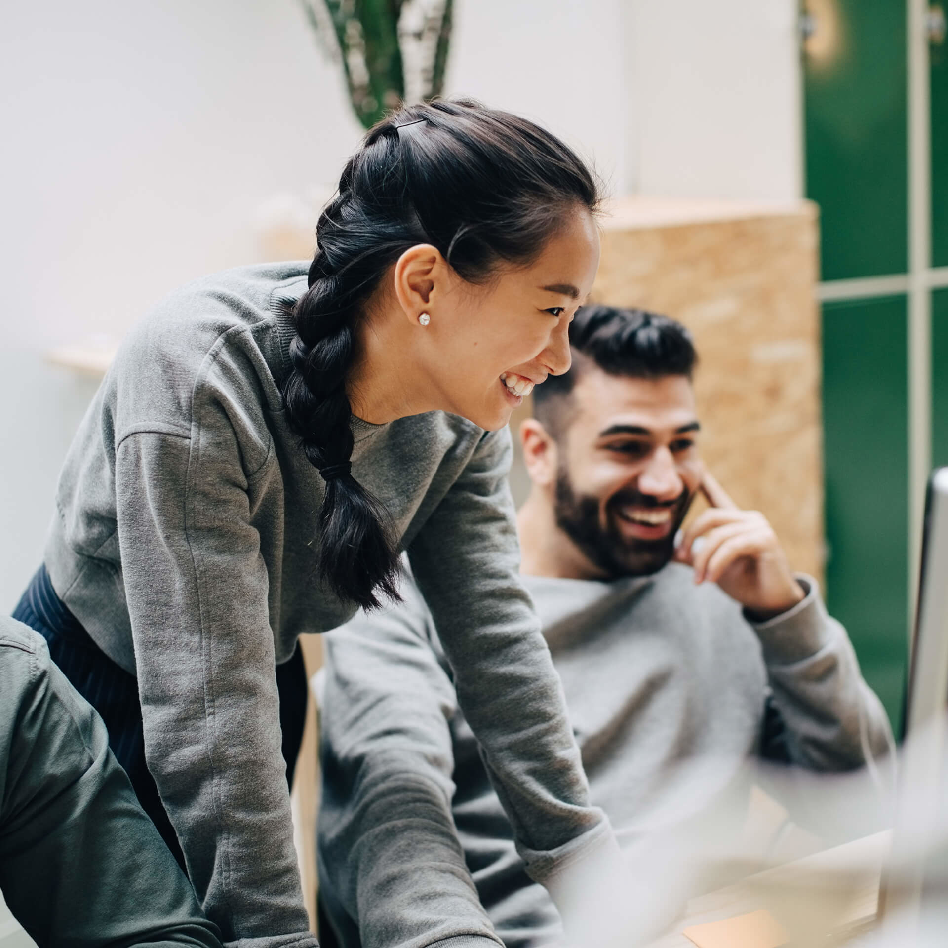 Two young professionals in casual grey sweaters share a genuine laugh while collaborating in a bright office space