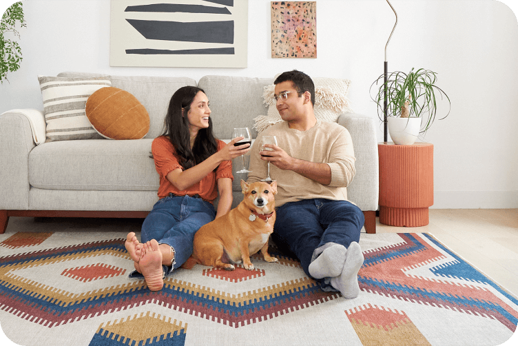 Couple relaxing on patterned rug with wine glasses and their ginger-colored dog, modern living room with abstract wall art