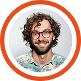 A smiling person with curly dark hair and glasses in a light blue shirt against an orange circular background